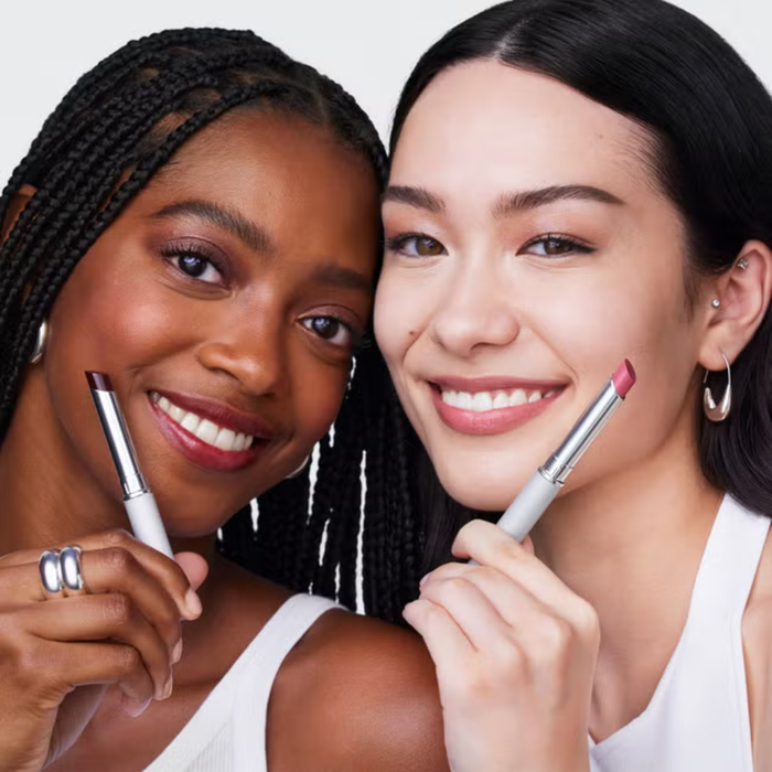 Two smiling models holding Clinique Cult Classic Lipstick Duo gift set lipsticks in white packaging against a light grey background.