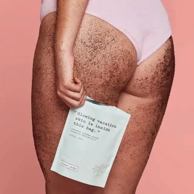 Frank Body Coconut Coffee Scrub pouch held against coffee scrub-covered legs on pink background, text reads "Glowing vacation skin is inside this bag."