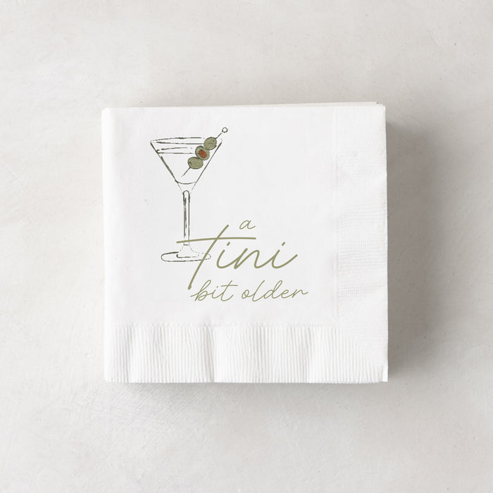 White 3-ply paper cocktail napkins with a martini glass and "a tini bit older" in olive green.