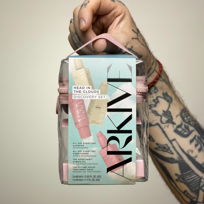 Tattooed hand holding Arkive Head In The Clouds Discovery Set in clear pink bag, sky-blue box on grey background.