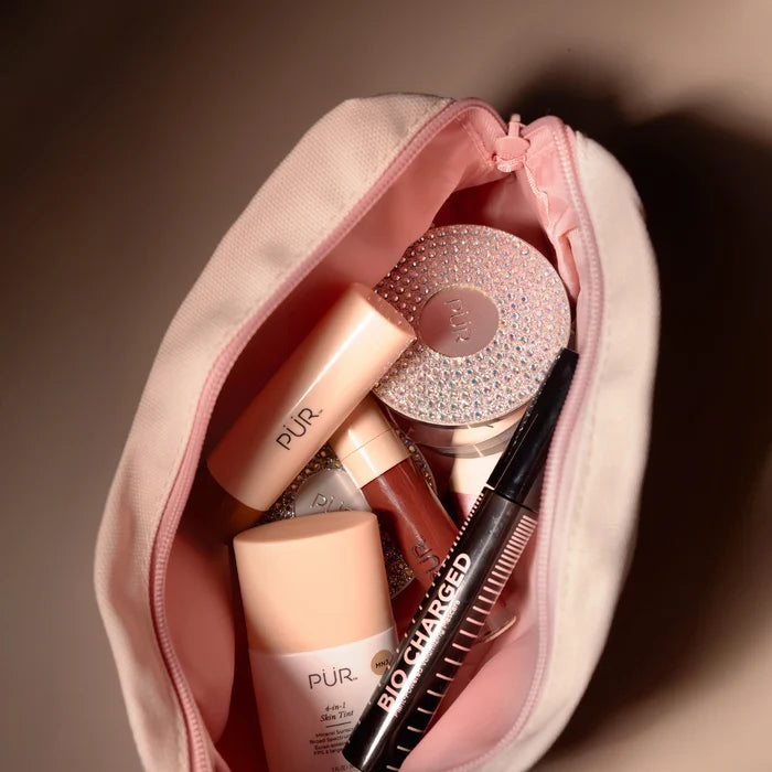 Open pink makeup bag filled with PUR cosmetics including a sparkling round compact, a pink bottle, lip gloss, and a black mascara.