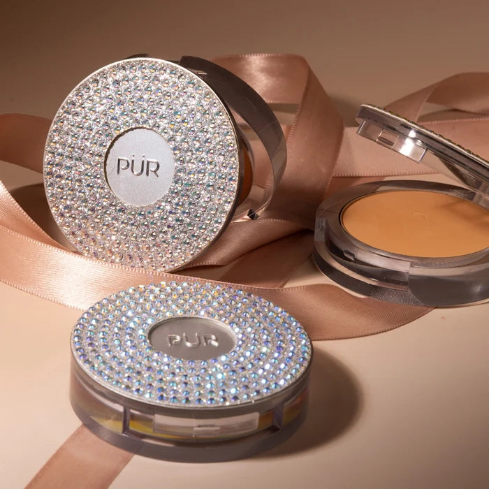 Round PUR pressed mineral makeup compact with sparkling rhinestone lid and beige powder visible, set against pink satin ribbon.