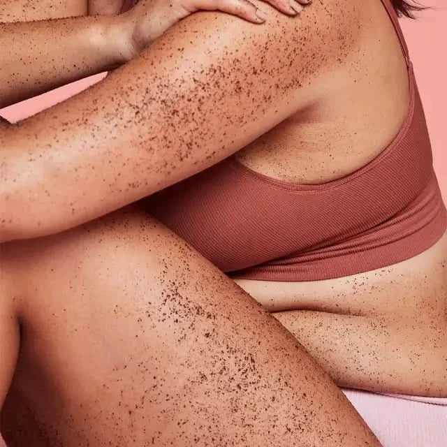 Frank Body Coconut Coffee Scrub applied on model's skin and legs, brown coffee granules visible, pink background.