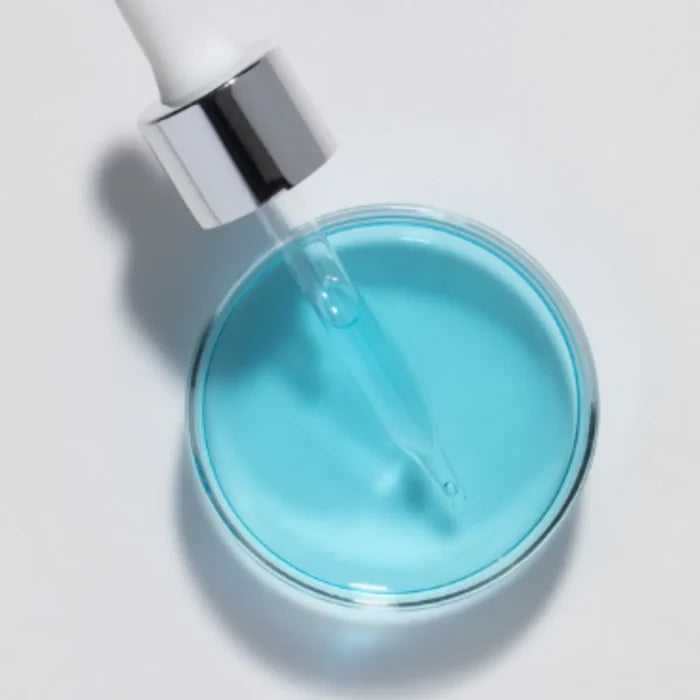 Dropper with silver collar dispensing blue serum into clear dish on white background
