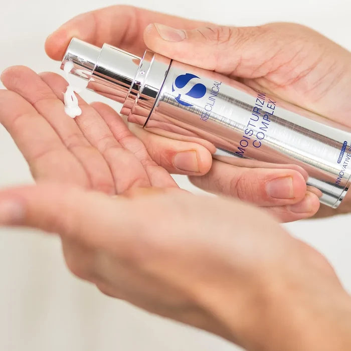 Hand dispensing white cream from silver pump bottle labelled iS Clinical Moisturizing Complex