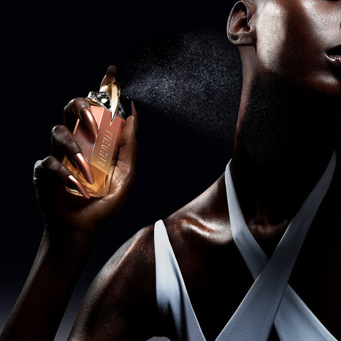 Mugler Alien Goddess Supra Florale EDP bottle with copper-rose casing held by a model spraying fragrance on black background.