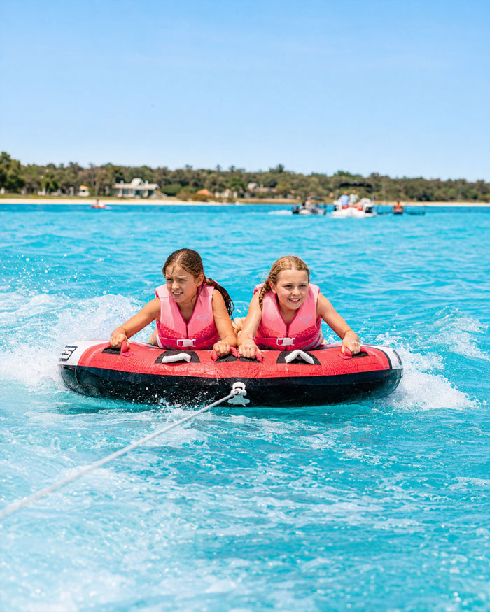 Two young girls wearing Roxy Youth Duraweave Vests, USCG approved pink life vests by Roxy, ride together on a red inflatable tube being pulled across bright blue water with a shoreline and trees in the background, ensuring fun and water safety.