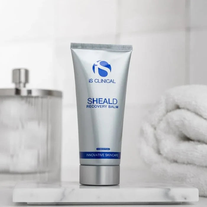 Silver tube of iS Clinical SHEALD Recovery Balm with blue text on marble surface, blurred towel and dispenser background