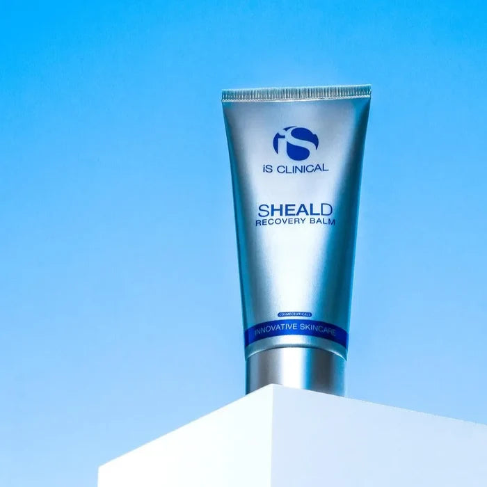 Silver tube of iS Clinical SHEALD Recovery Balm with blue label on white pedestal against blue background