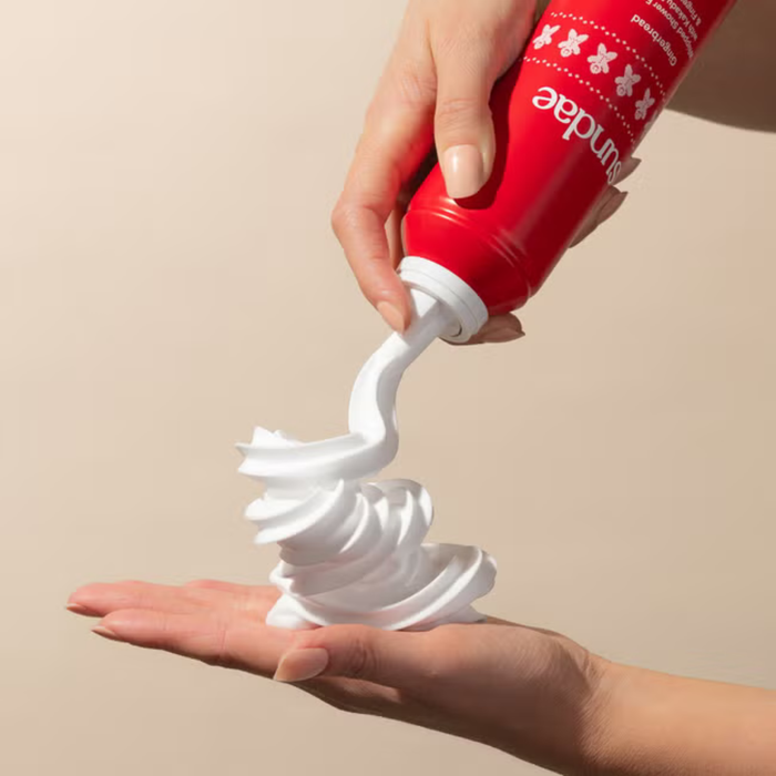 Sundae Gingerbread Shower Foam dispensed as white whipped swirl onto palm, red can with white text, beige background.