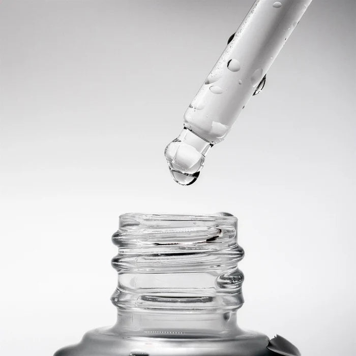 Glass dropper bottle with clear liquid being dispensed on a light gray background