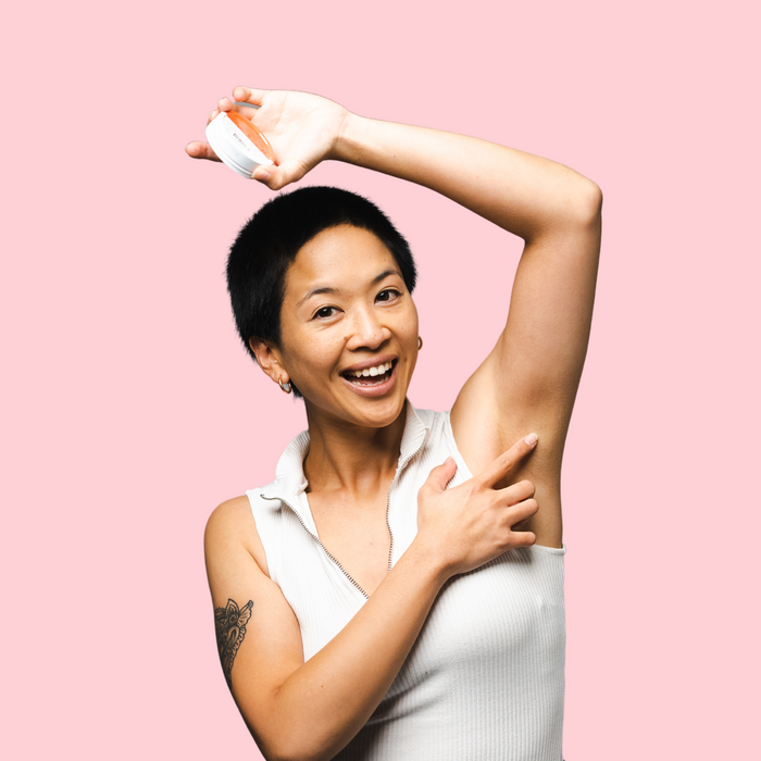 Woohoo Natural Deodorant Paste (Urban) in a person's hand, applied to the underarm against a pink background.