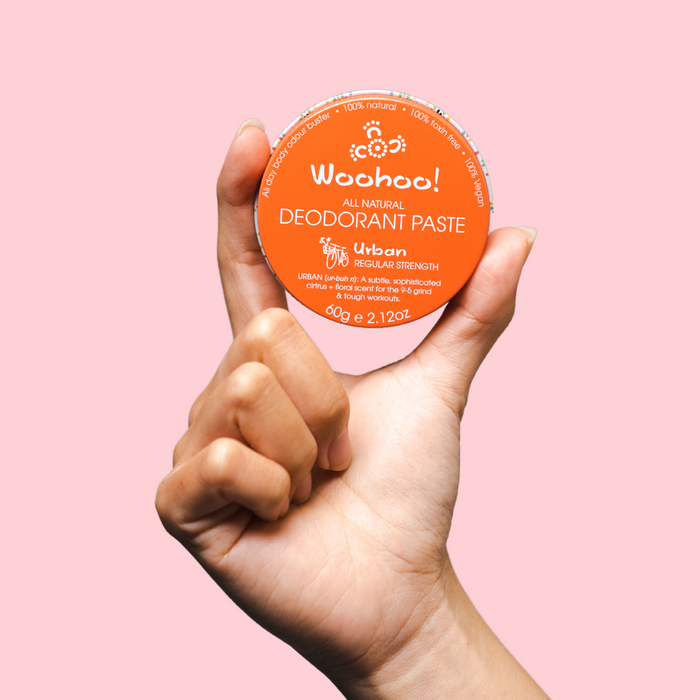Woohoo Natural Deodorant Paste (Urban) in a hand, featuring an orange container with white text on a pink background.