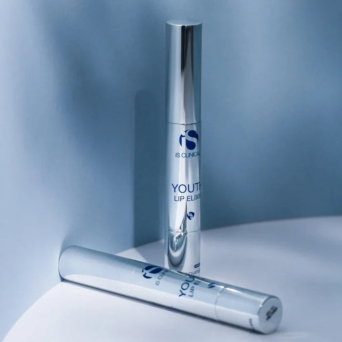 Two silver cylindrical tubes of iS Clinical Youth Lip Elixir on a curved white and blue surface